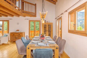Comedor con mesa de madera dispuesta para seis personas, rodeada de sillas. Se observa un aparador de madera con puertas de cristal, un reloj en la pared y vistas al exterior con vegetación.