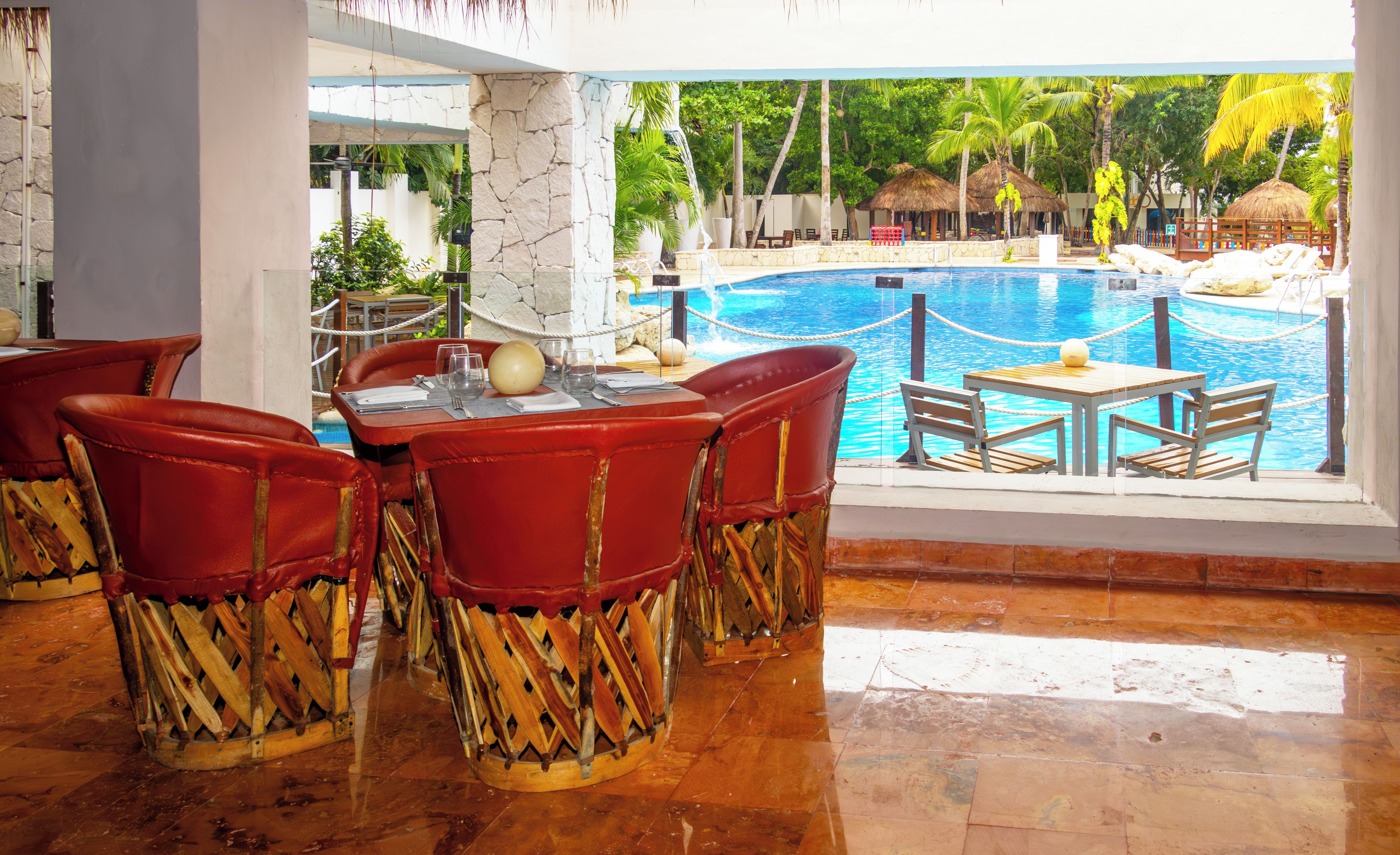 A poolside dining area with red chairs and a table, overlooking a swimming pool and tropical scenery.