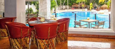 A poolside dining area with red chairs and a table, overlooking a swimming pool and tropical scenery.