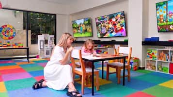 Una habitación con dos niños jugando videojuegos, un tapete de juego colorido, y un área de juegos infantil con juguetes y una cocinita.