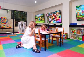 Una habitación con dos niños jugando videojuegos, un tapete de juego colorido, y un área de juegos infantil con juguetes y una cocinita.