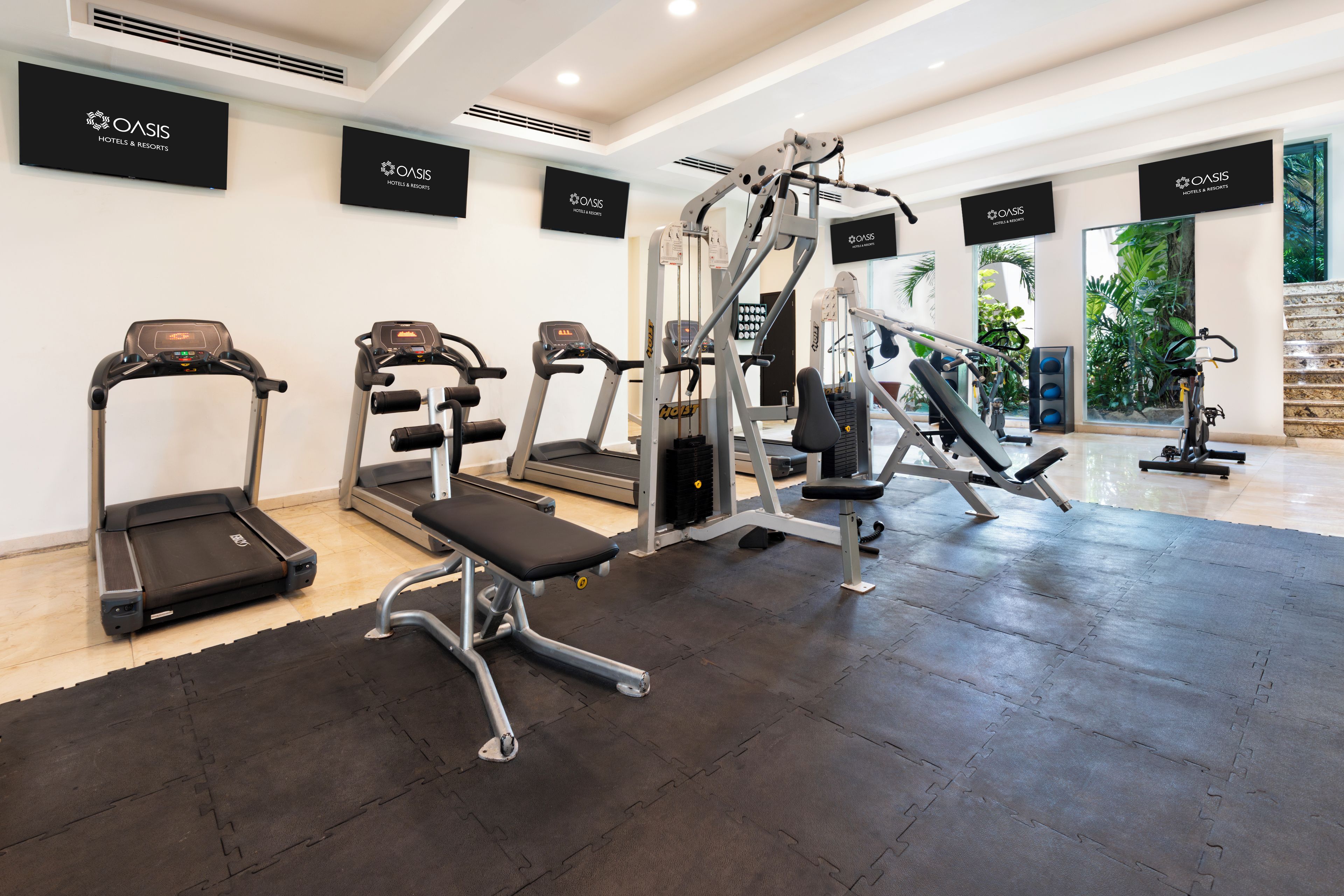 Un gimnasio con cintas de correr, una prensa de banca y máquinas de pesas, con letreros que dicen 'OASIS HOTELS & RESORTS' en las paredes.