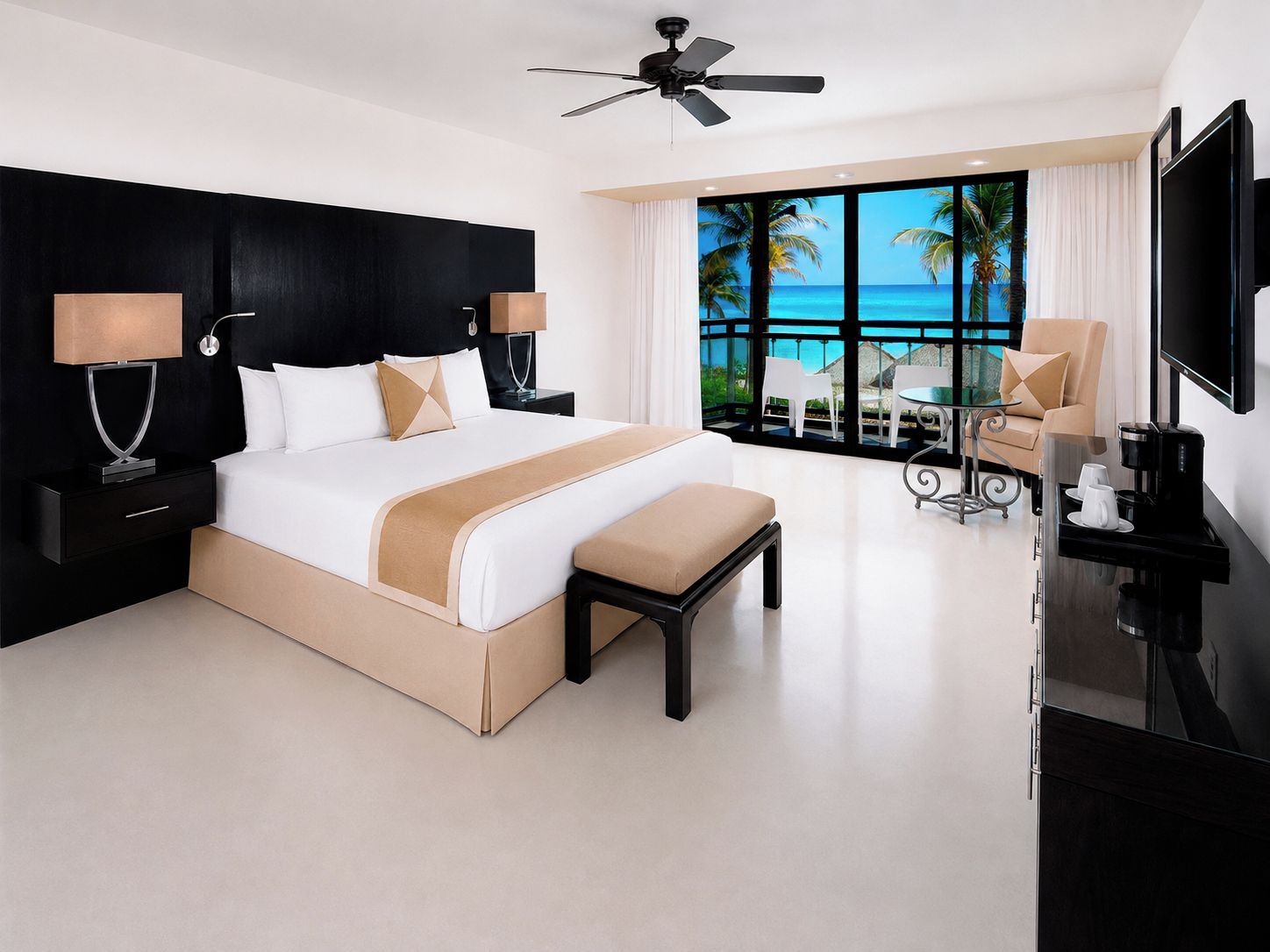 Una habitación de hotel moderna con una cama grande, ventilador de techo, televisión y balcón con vista al océano.