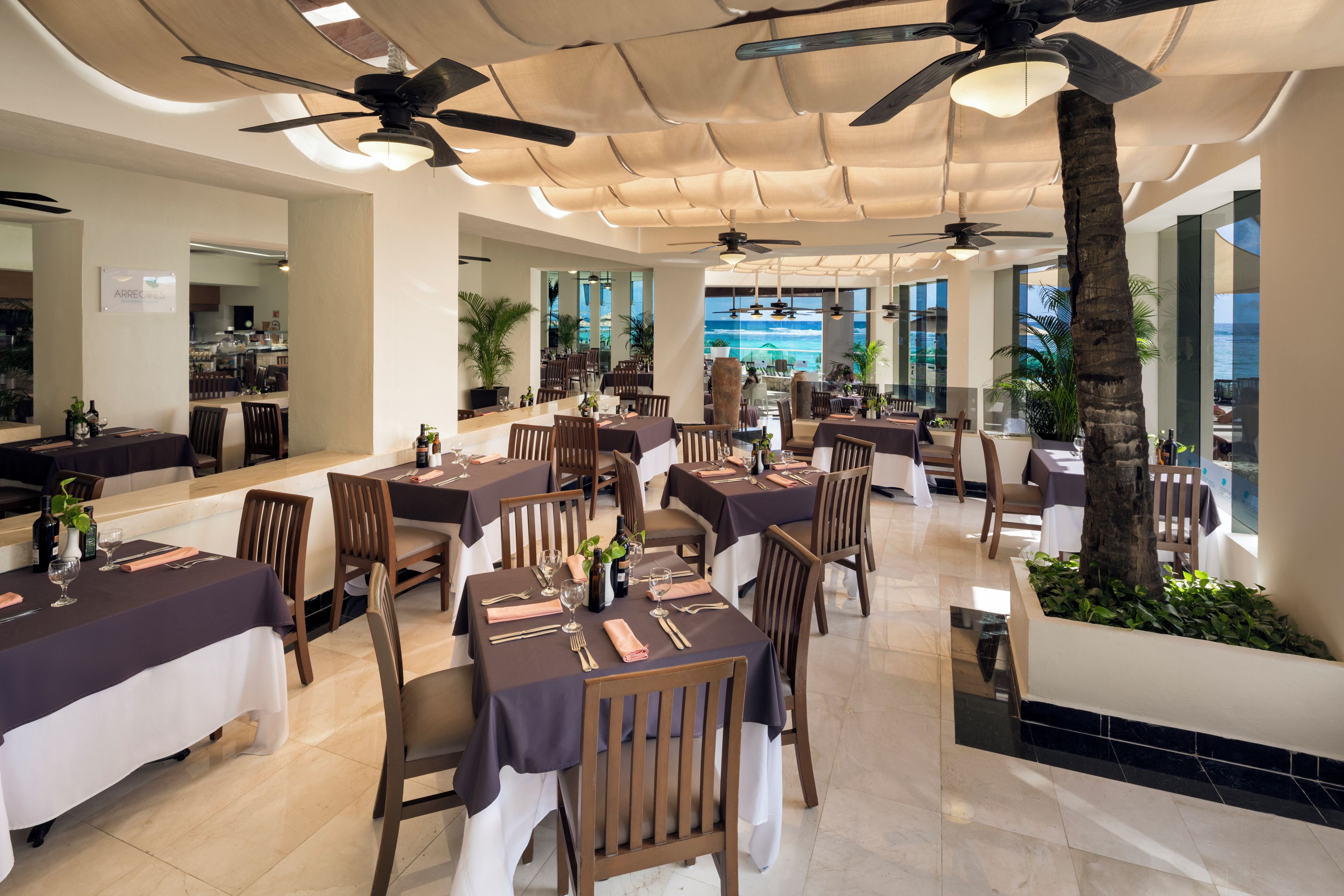 A restaurant with multiple tables, ceiling fans, and a view of the ocean.