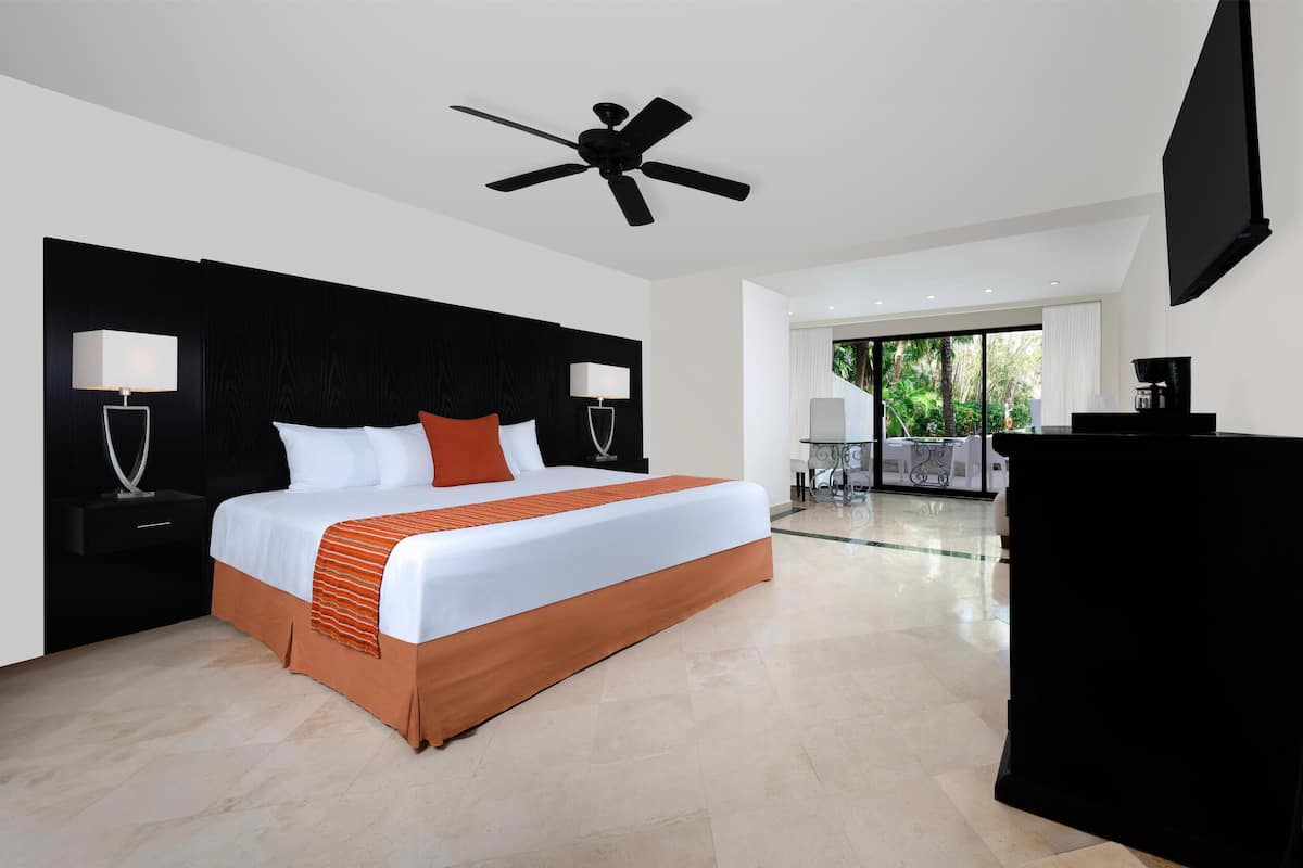 A modern hotel room with a large bed, a ceiling fan, a TV, and a balcony with a view of greenery.