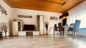 A modern living room with a wooden ceiling, a flat-screen TV, a dining area with chairs, and a vase with dried plants. - Gemütlicher Bungalow in Ruhiger Lage mit Traumhafter Aussicht für 2-6 Personen (Dipperz)