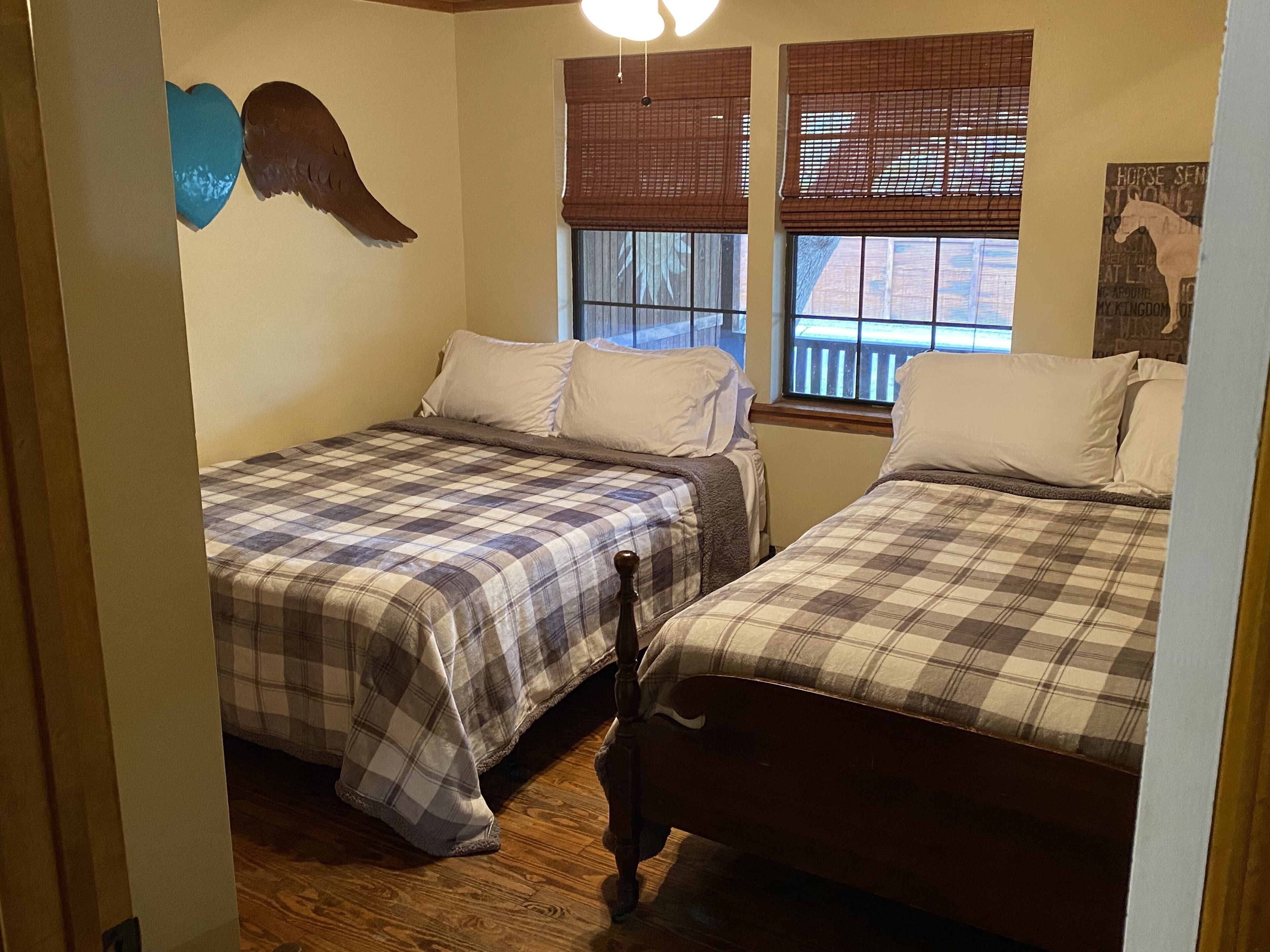 Un dormitorio con dos camas, cada una con cubrecama de cuadros, cabeceros de madera y almohadas blancas. Hay dos ventanas con persianas marrones, y una lámpara fijada a la pared.
