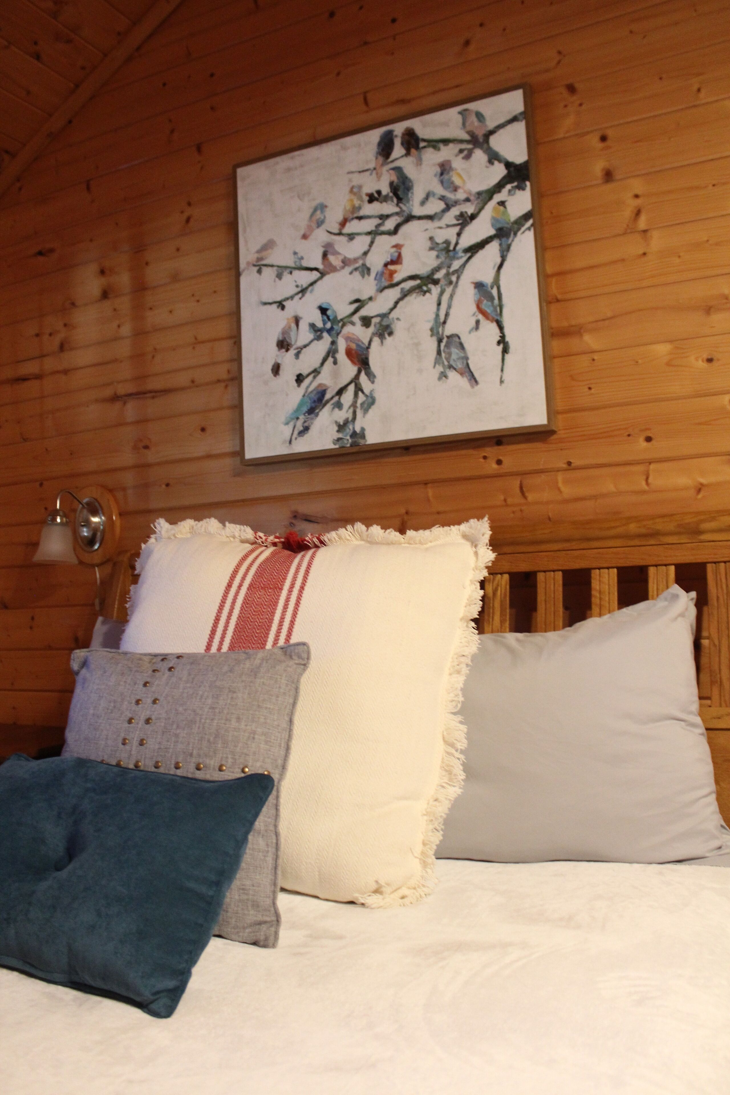 A wooden cabin bedroom with a bed, pillows, a wall-mounted lamp, and a painting of birds on a branch.