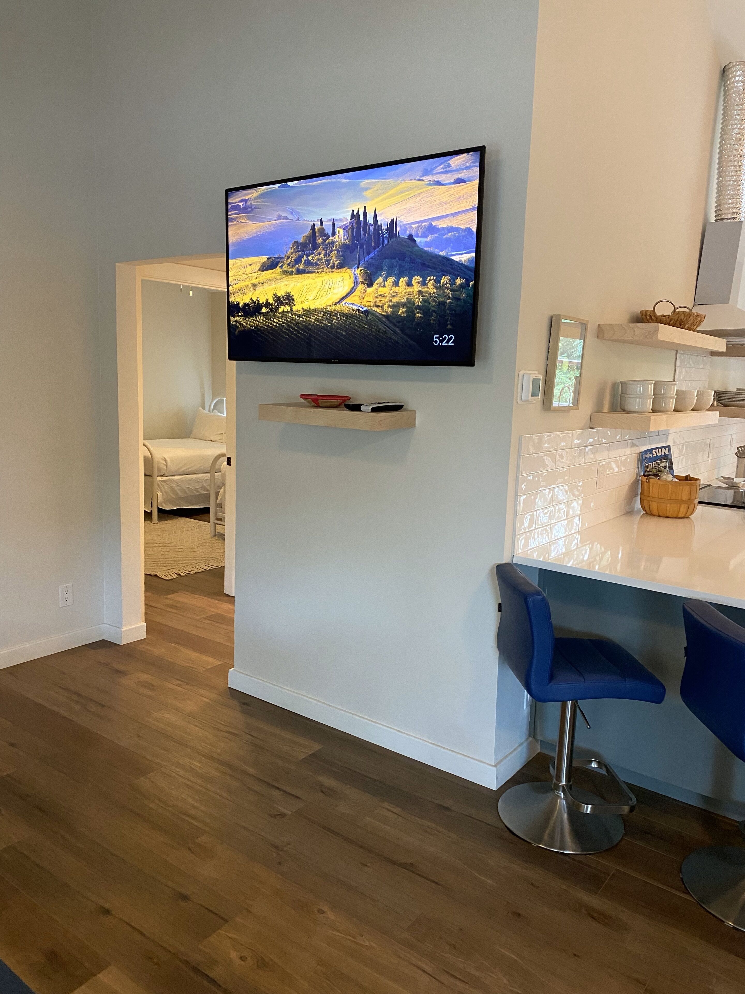 A modern living room with a flat-screen TV mounted on a wall, wooden flooring, a blue bar stool, and a kitchen area with white cabinets and a countertop.