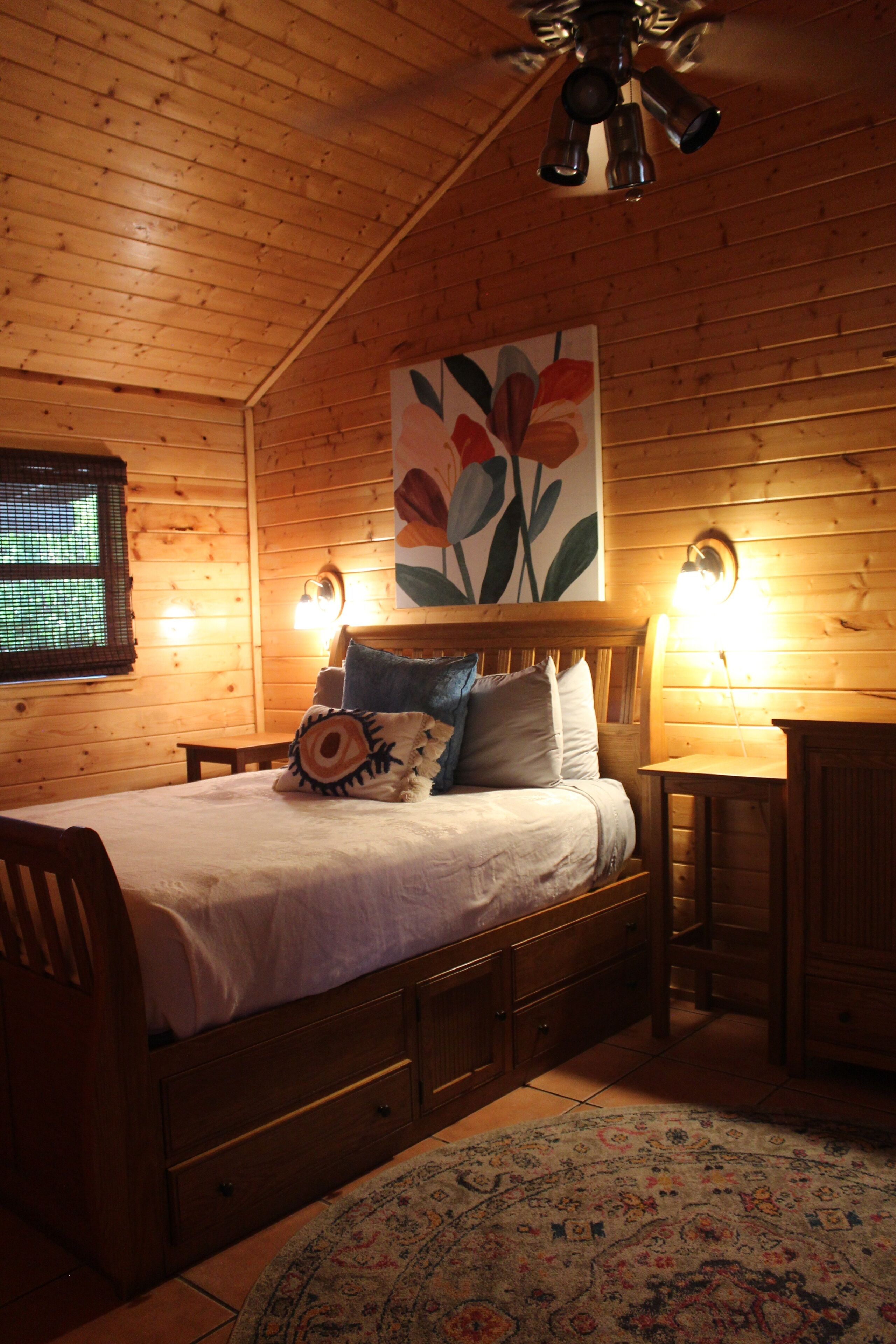 A wooden cabin bedroom with a bed, pillows, a nightstand, a ceiling fan, and a wall-mounted lamp.