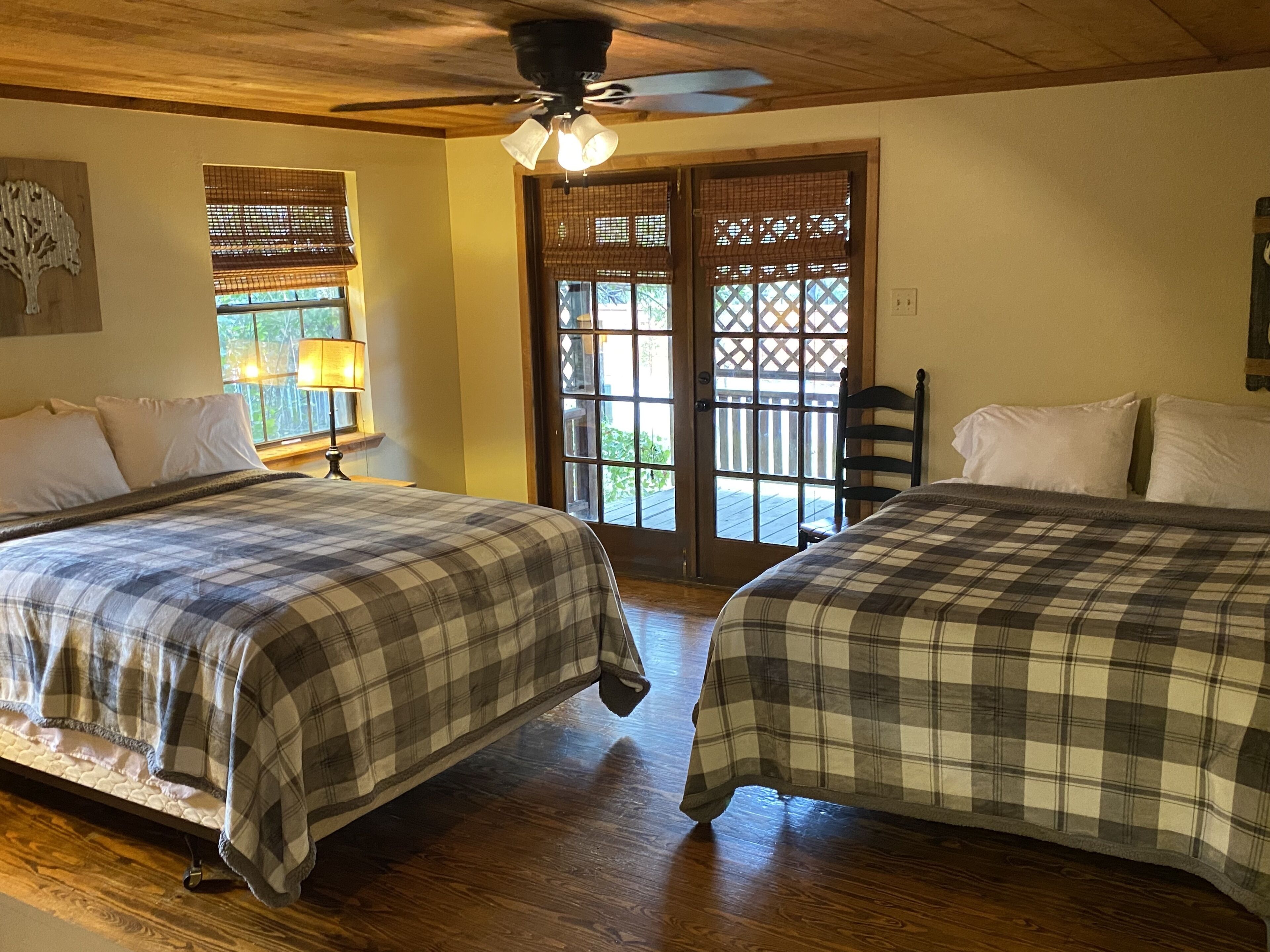 A room with two beds, wooden flooring, a ceiling fan, and a door leading to an outdoor area.