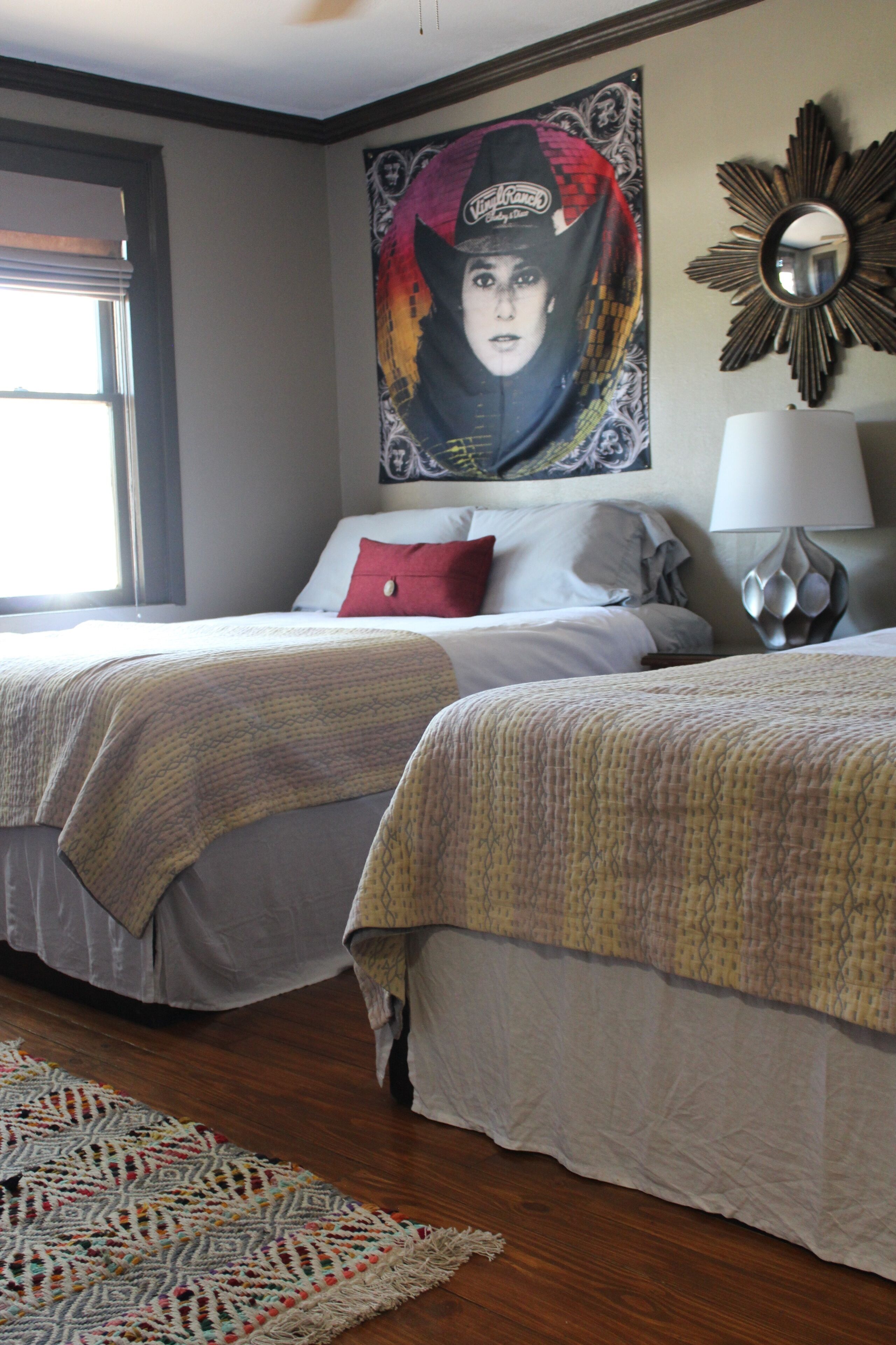 A hotel room with two beds, a large poster on the wall, a mirror, and a lamp.