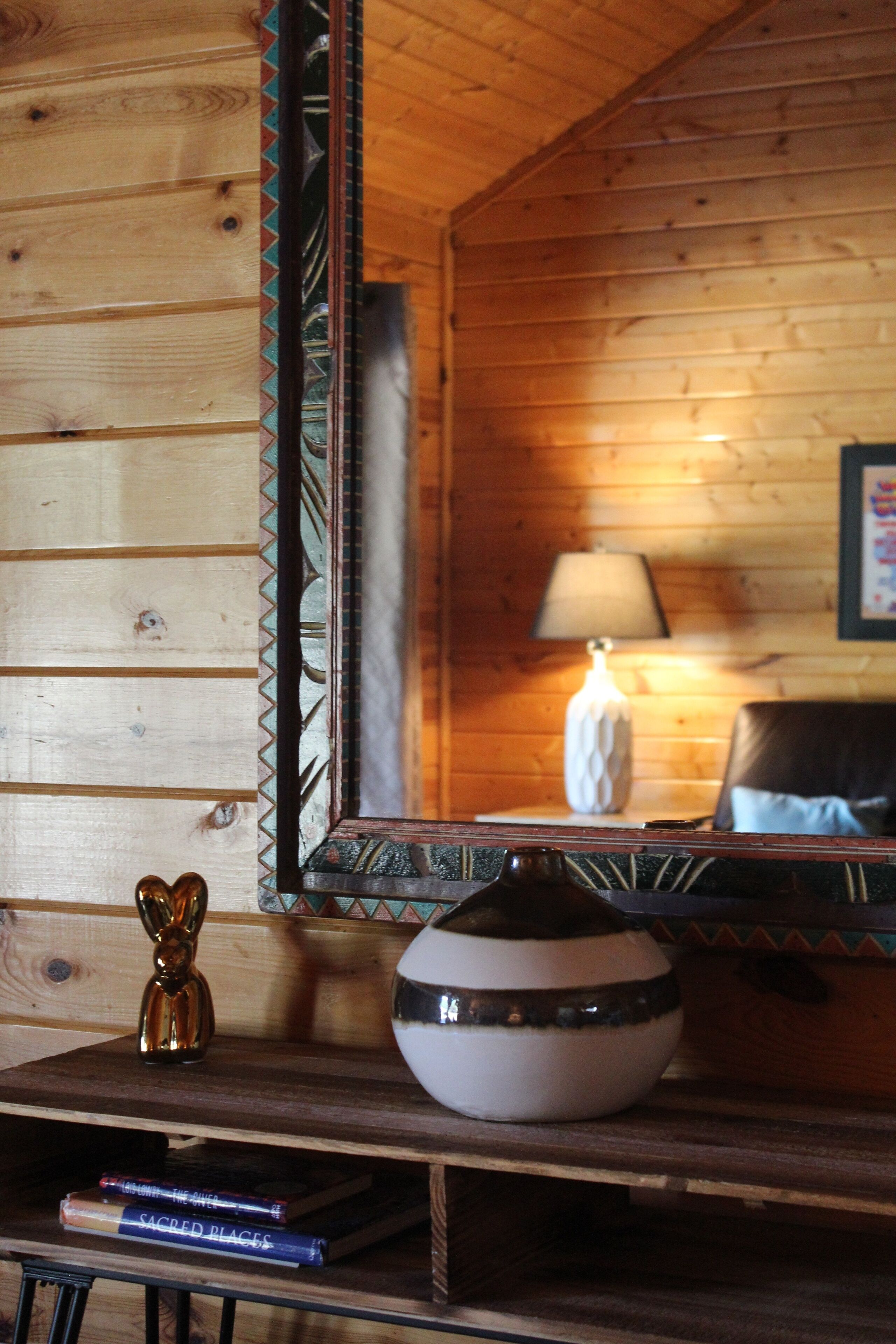Un interior de madera que muestra un espejo reflejando una lámpara, una silla, una figurita y un cuenco sobre una estantería.