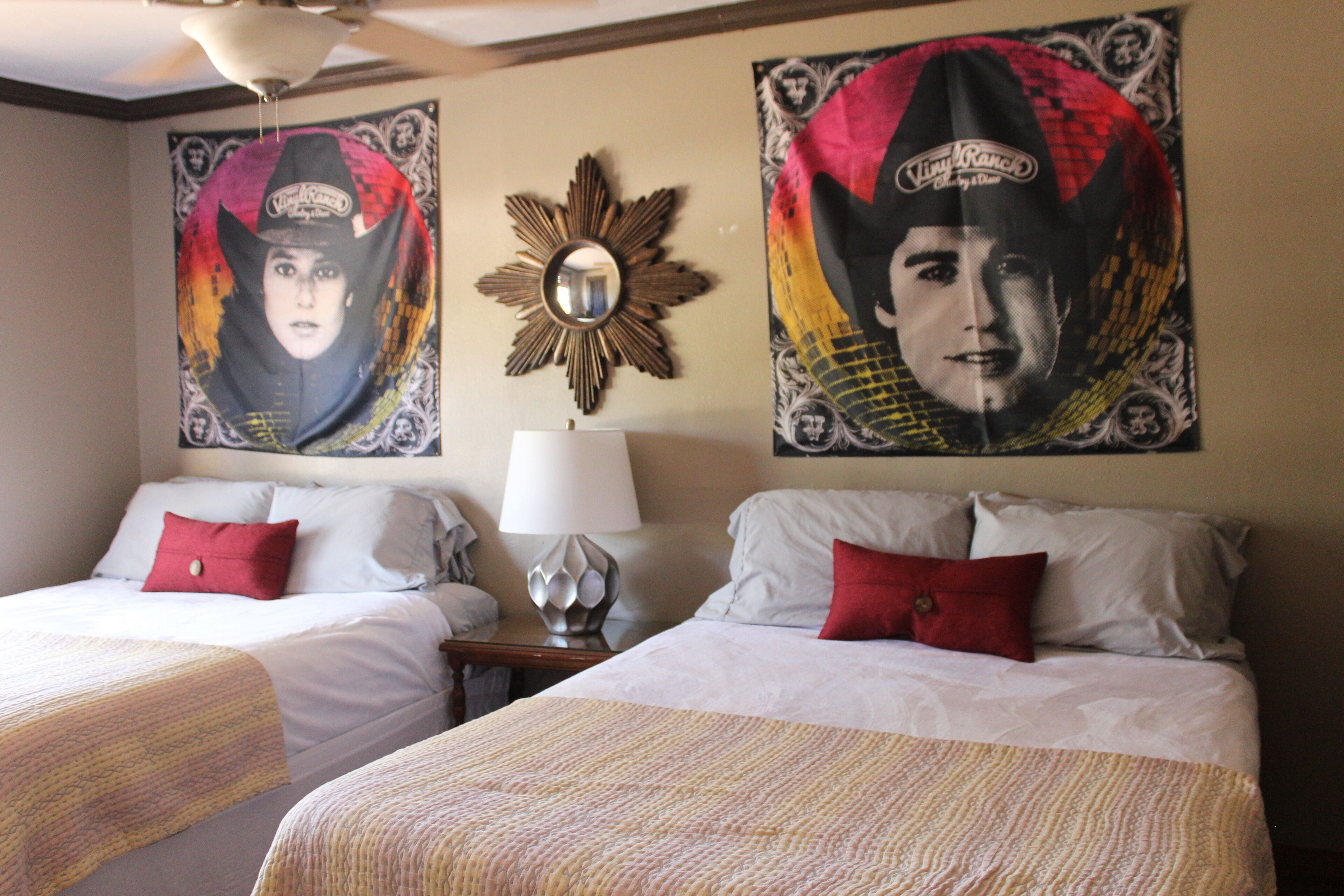 Une chambre d’hôtel avec deux lits, chacun agrémenté de draps blancs et d’oreillers rouges, une table de chevet avec une lampe et deux grandes affiches au mur.