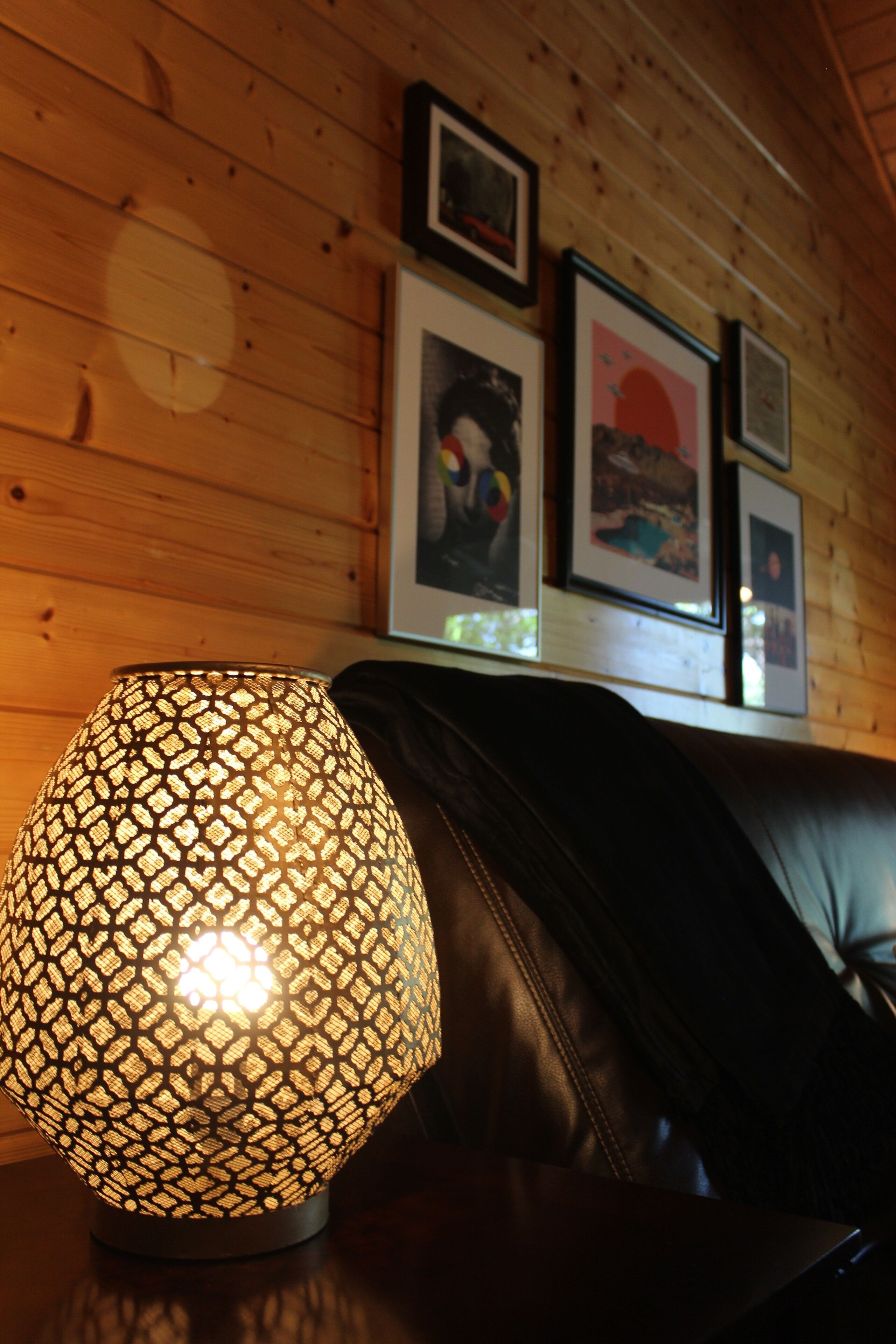 A lamp with a patterned shade on a table, a leather couch, and framed pictures on the wall.