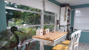 A dining area with a wooden table and white chairs, a window with a view of greenery, and a birdhouse on the window sill. - Casa Las Palmeras (Port Richey)