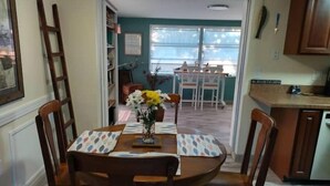 A dining area with a wooden table, chairs, and a vase with flowers. There is a kitchen area with cabinets and a countertop visible in the background. - Casa Las Palmeras (Port Richey)