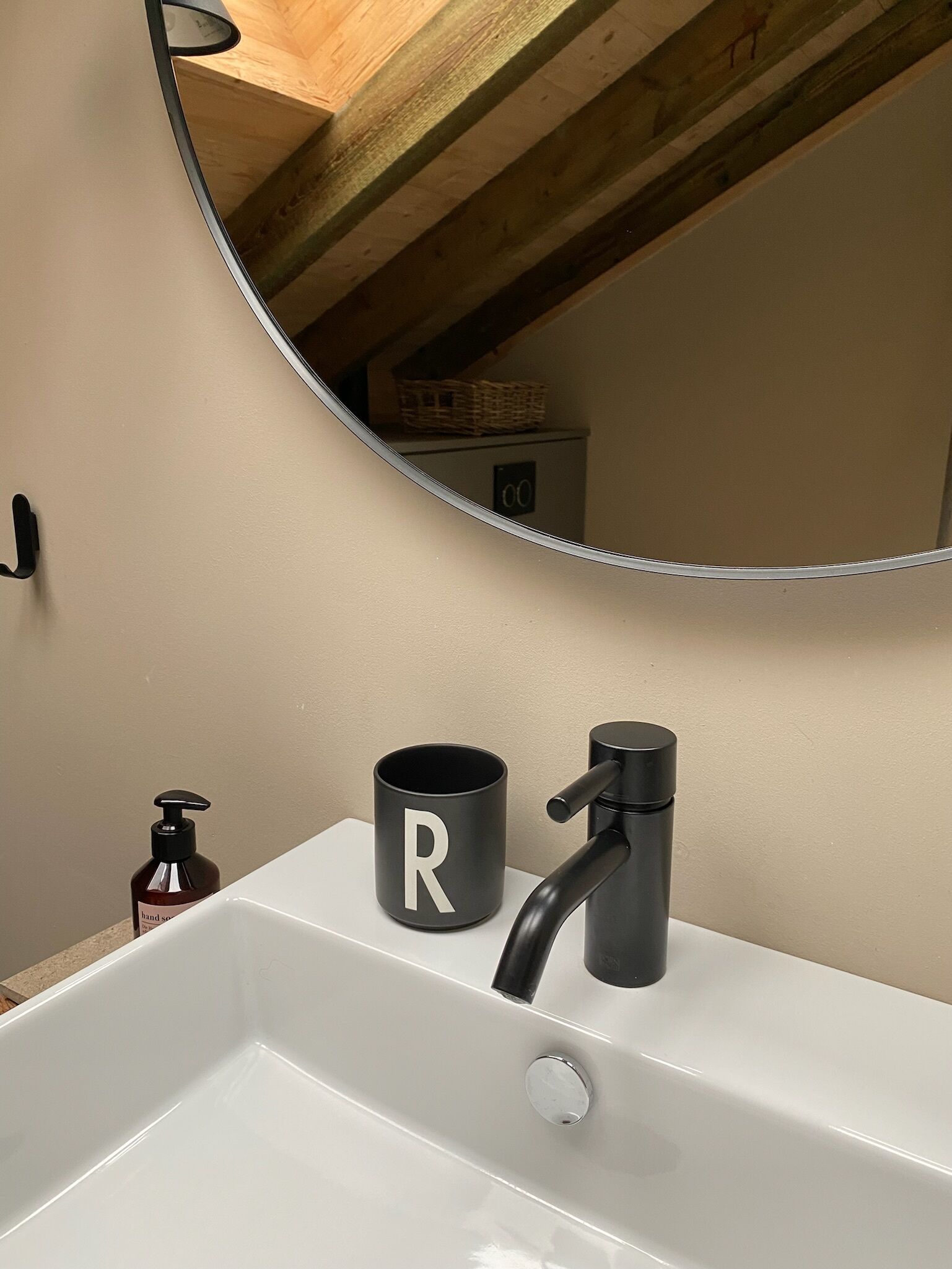 Ein Badezimmer mit einem weißen Waschbecken, einem schwarzen Wasserhahn, einer schwarzen Tasse mit einem weißen 'R' und einer Seifenspender.