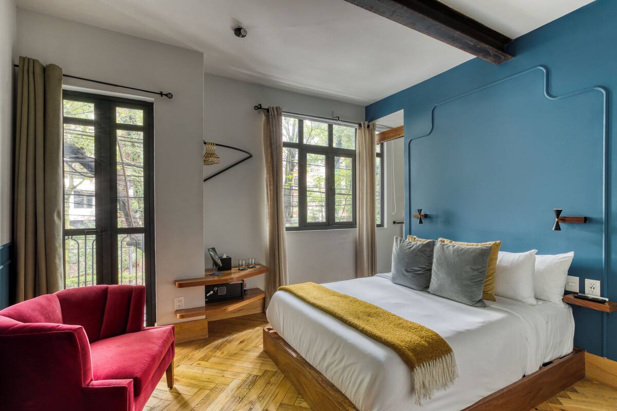 Un dormitorio con una cama, una silla roja, una mesita de noche de madera y una ventana con cortinas.
