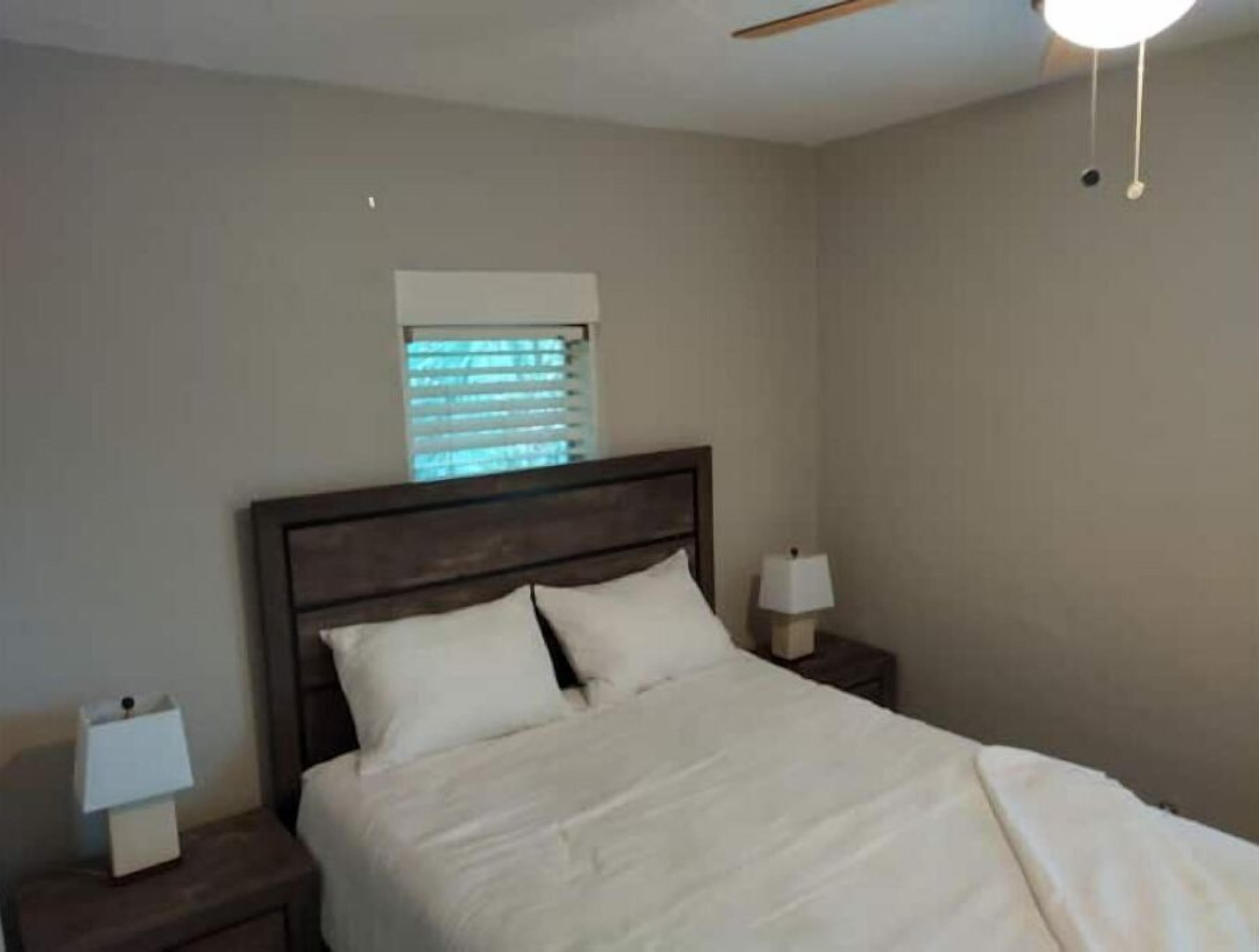 A bedroom with a bed, two bedside tables with lamps, a window with blinds, and a ceiling fan.