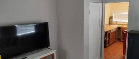 Une pièce dotée d’un téléviseur à écran plat posé sur un meuble blanc, d’un ventilateur de plafond et d’un coin cuisine avec une fenêtre et un comptoir.