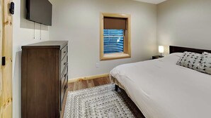 A bedroom with a bed, a dresser, a TV mounted on the wall, a window with blinds, and a nightstand with a lamp. - Moon and Stars by Taylor Made Deep Creek (McHenry)