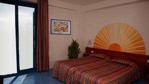 Camera d'albergo con un letto, un'ampia finestra con tende e un quadro appeso al muro, oltre a un motivo solare sulla parete.