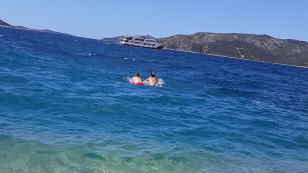 A beach with clear shallow waters, a boat in the distance, and rocky terrain in the background.