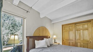 Ein Schlafzimmer mit Bett, Fenster mit Blick auf Bäume, Holz-Kopfteil, Nachttisch mit Lampe und Holztür.