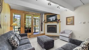 Ein Wohnzimmer mit Kamin, einem darüber angebrachten Fernseher und einem großen Fenster mit Blick auf Bäume. Zwei Sofas und ein Sessel sind ebenfalls vorhanden.