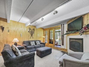 A cozy living room with a fireplace, wooden walls, and a large window.