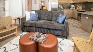 A modern living room with a grey sofa, two ottomans, a wooden bench, and a small table. A kitchen with wooden cabinets and a refrigerator is visible in the background. - Cozy Vail Retreat, On Shuttle Route, BBQ (Vail)