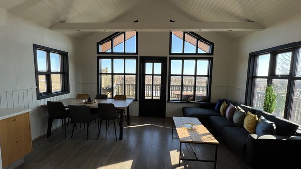 A room with a dining table, chairs, a sofa, and large windows offering views of the outdoors. - Minni-Borgir Cabins (Grímsnes- og Grafningshreppur)