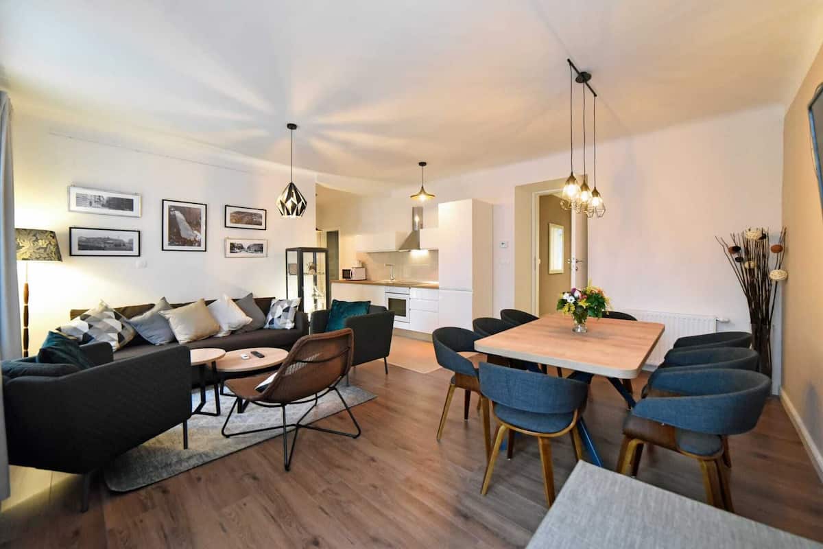 A modern living room with a sofa, chairs, and a dining table. There is a kitchen in the background.