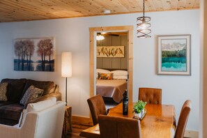 A cozy living room with a wooden ceiling, a dining table with chairs, a sofa, a lamp, and artwork on the walls.
