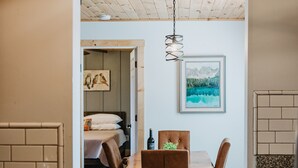 A wooden dining table with chairs, a kitchen area with a wooden floor, and a bedroom with a bed and a painting.