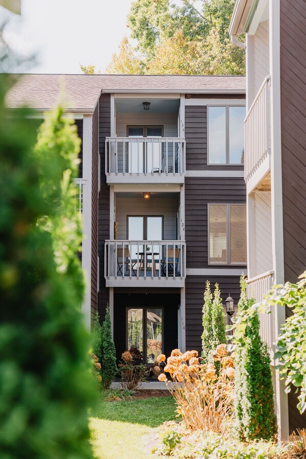 Un bâtiment à deux étages doté d’un balcon au deuxième étage, d’un espace extérieur couvert aménagé en coin salon et d’un jardin bien entretenu où poussent diverses plantes et fleurs.