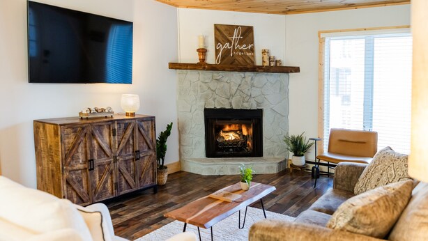 A living room with a stone fireplace, a mounted TV, and a 'gather together' sign above the fireplace.