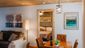 A cozy living room with a wooden ceiling, a dining table with chairs, a sofa, a lamp, and artwork on the walls.