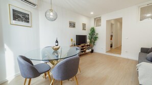 Una sala de estar moderna con una mesa de comedor de vidrio, sillas tapizadas de color gris, un televisor de pantalla plana, una planta en maceta y un sofá.
