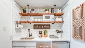 A compact kitchen with white cabinets, a stainless steel dishwasher, and a microwave. There are wooden shelves above the sink holding dishes and a sign that says "cook. sing. dance."