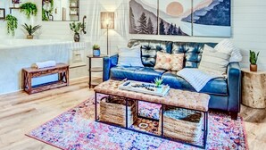 A cozy living room with a blue sofa, a coffee table, and a colorful rug. There are plants on shelves and a painting on the wall.