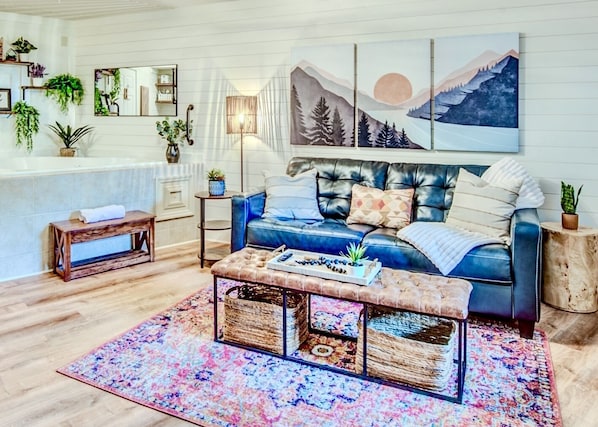 A cozy living room with a blue sofa, a coffee table, and a colorful rug. There are plants on shelves and a painting on the wall.
