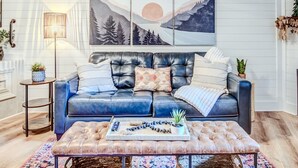 A modern living room with a blue leather sofa, a tufted ottoman, and a large wall-mounted painting depicting a mountain landscape.