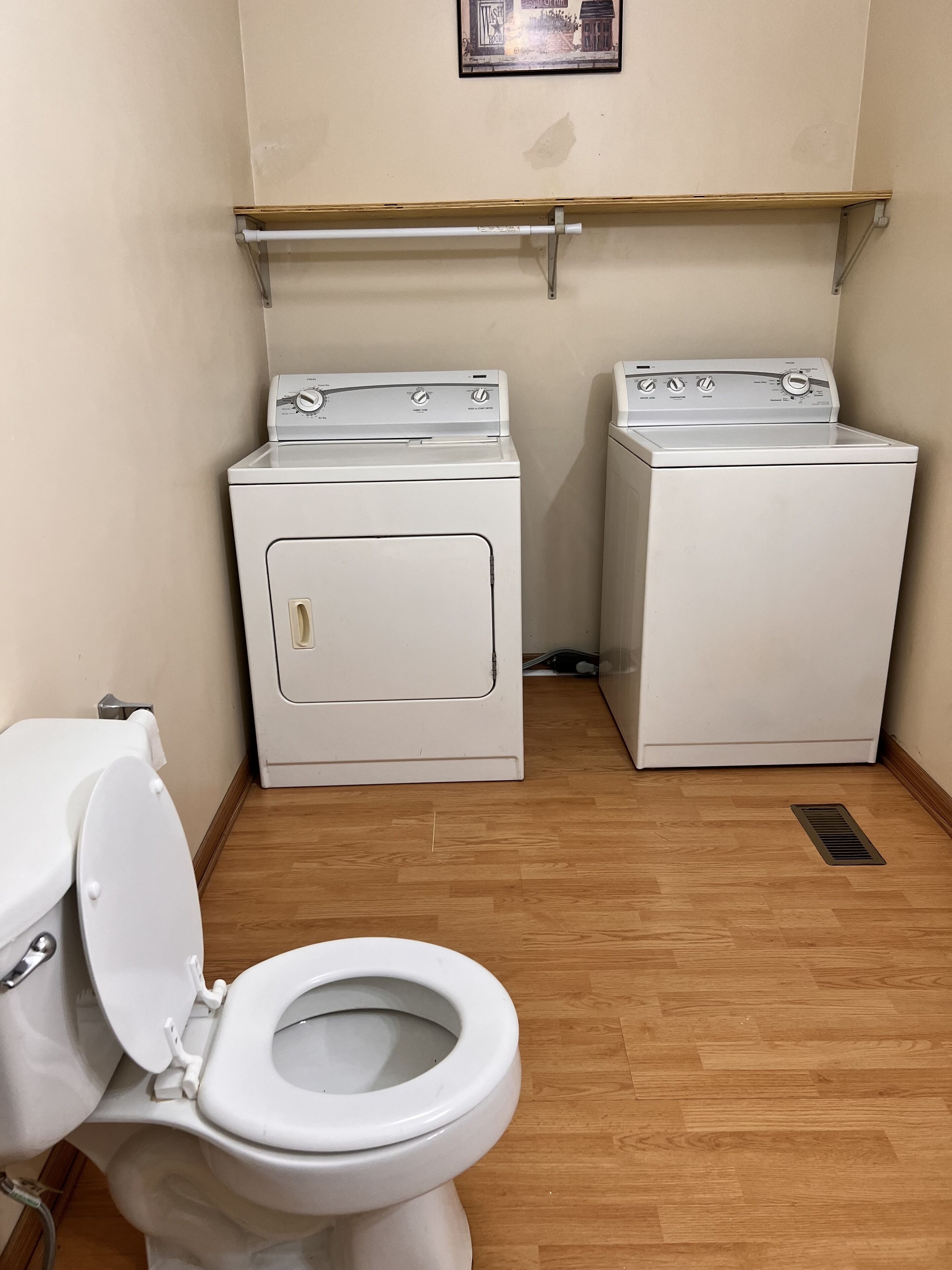 Una petita sala de bugaderia amb dues rentadores blanques, un vàter i un terra de fusta.