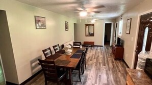 Un comedor amplio con una larga mesa de madera y sillas, un ventilador de techo y un pasillo que conduce a otras habitaciones.