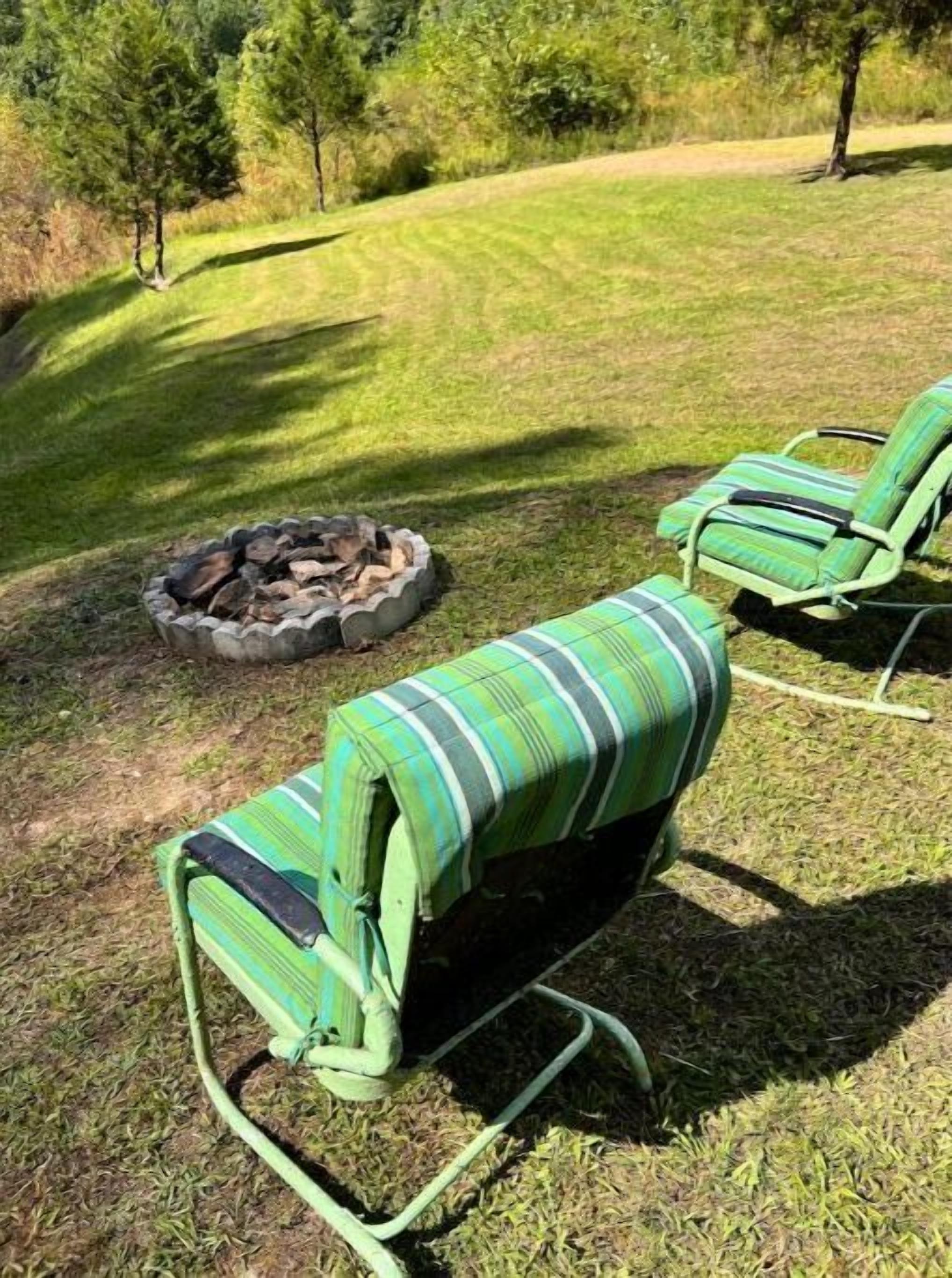 Twee groene ligstoelen met streepkussens, een vuurkuil en een gazon met bomen op de achtergrond.