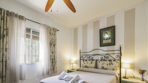 Un dormitorio con cama, dos mesitas de noche, una mesita auxiliar, una ventana con cortinas, un ventilador de techo y un cuadro en la pared.