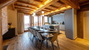 Un espace repas avec une longue table dressée pour six personnes, des poutres en bois au plafond et une cuisine en arrière-plan.
