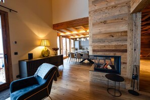 A modern living room with a fireplace, wooden ceiling beams, a dining area, and a sofa. - Goûtez l'élégance de ce chalet d'exception. (Peisey-Nancroix)